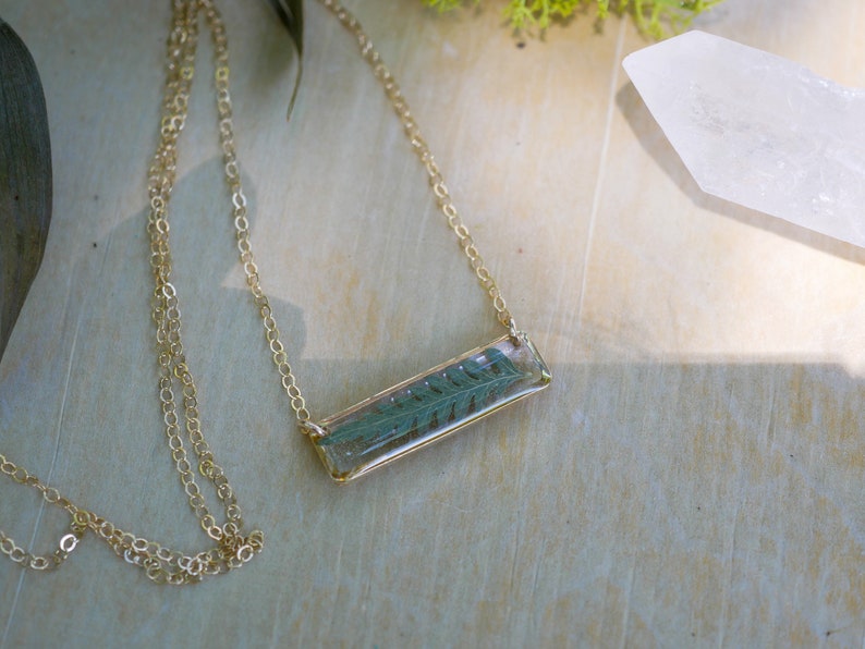 May include: A gold chain necklace with a rectangular pendant featuring a pressed fern leaf encased in clear resin.