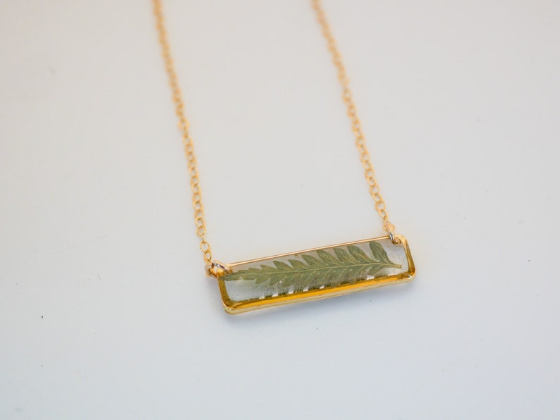 May include: A gold chain necklace with a rectangular pendant featuring a pressed fern encased in clear resin.
