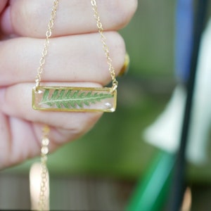 May include: A gold chain necklace with a rectangular pendant featuring a pressed fern in clear resin.