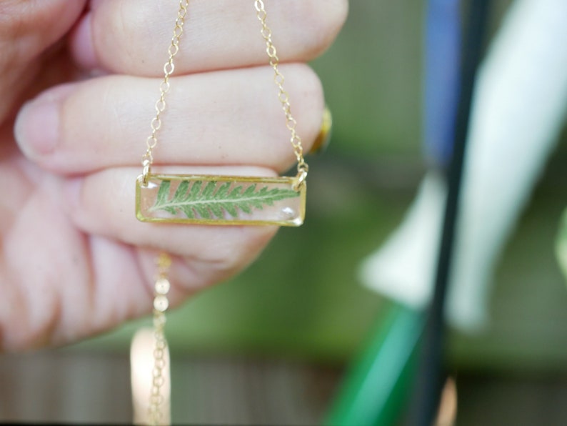May include: A gold chain necklace with a rectangular pendant featuring a pressed fern in clear resin.