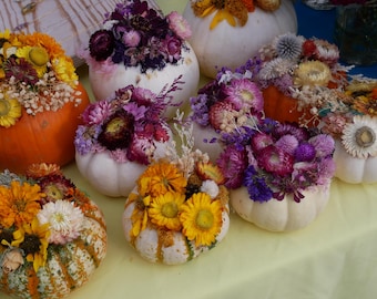 May include: A collection of white pumpkins decorated with colorful dried flowers. The pumpkins are arranged on a light yellow surface.