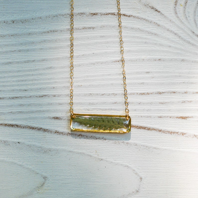 May include: A gold chain necklace with a rectangular pendant featuring a pressed green leaf encased in clear resin.