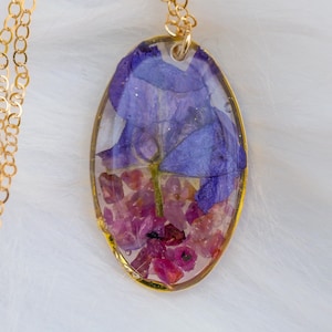 May include: A gold-colored chain necklace with a pendant. The pendant is an oval shape with a gold border, encasing dried purple and pink flowers in clear resin. The necklace is on a white, fluffy surface.
