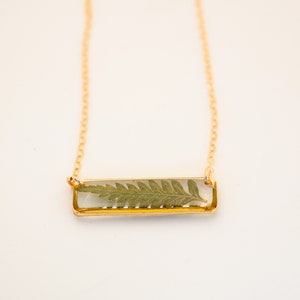 May include: A gold chain necklace with a rectangular pendant featuring a pressed fern leaf encased in clear resin.