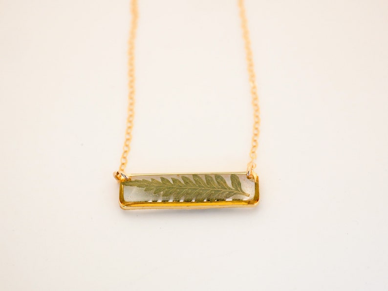 May include: A gold chain necklace with a rectangular pendant featuring a pressed fern leaf encased in clear resin.