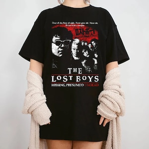 May include: Black t-shirt with a graphic from the film "The Lost Boys". The design includes character images and the text "THE LOST BOYS MISSING, PRESUMED UNDEAD". Additional text at the top reads "Sleep all day. Party all night. Never grow old. Never die. It's fun to be a vampire."