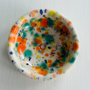 May include: A small, round ceramic bowl with a white base featuring a vibrant, multi-coloured splatter pattern. The design includes splashes of bright orange, teal, yellow, and dark blue paint across the interior and rim, with a small hole at the centre.