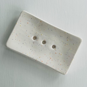 May include: A speckled ceramic soap dish. The white dish has small black, orange, and yellow speckles. It features three drainage holes and a gently curved design. Measurements are not available.
