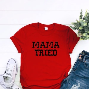 K&ouml;nnte beinhalten: Rotes T-Shirt mit schwarzem Text, der "MAMA TRIED" lautet. Das Shirt liegt auf einer wei&szlig;en Holzoberfl&auml;che mit einem Paar wei&szlig;en Turnschuhen und einer blauen Jeans.