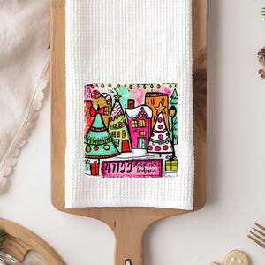May include: White waffle-weave kitchen towel with a colourful Christmas-themed graphic. The graphic features decorated trees, houses, and the text "47122, Keigstown, Indiana". The towel is displayed on a wooden cutting board.
