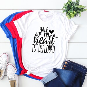 K&ouml;nnte beinhalten: Ein wei&szlig;es T-Shirt mit dem Text "HALF OF MY HEART IS DEPLOYED" in schwarzer Schrift, einem schwarzen Herz und einem blau, rot und wei&szlig;en Farbschema. Das Shirt wird mit blauen Jeans und wei&szlig;en Turnschuhen kombiniert. Eine kleine gr&uuml;ne Pflanze ist im Hintergrund.