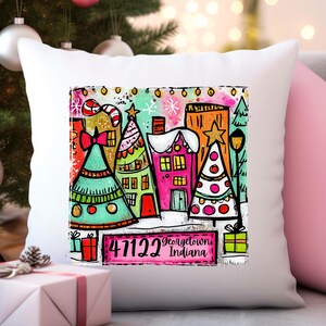 May include: White decorative pillow featuring a colorful Christmas village scene. The design includes decorated trees, houses, and the text "47122 Georgetown Indiana". The pillow has a festive, whimsical aesthetic.