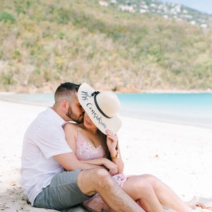 May include: A couple sitting on a white sandy beach, the woman is wearing a straw hat with the text "Mrs. Considine" written on it.