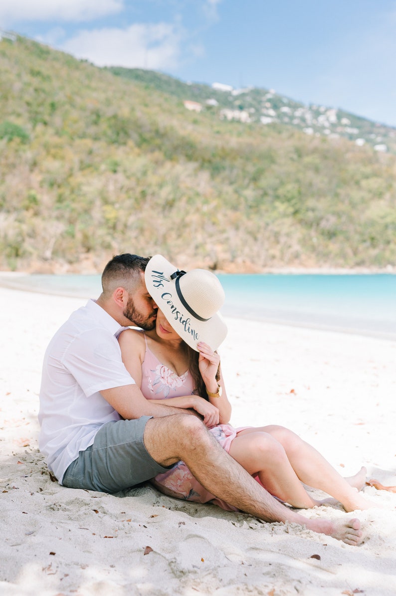 May include: A couple sitting on a white sandy beach, the woman is wearing a straw hat with the text "Mrs. Considine" written on it.