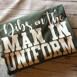 May include: A green t-shirt with white text that reads "Dibs on the man in uniform".