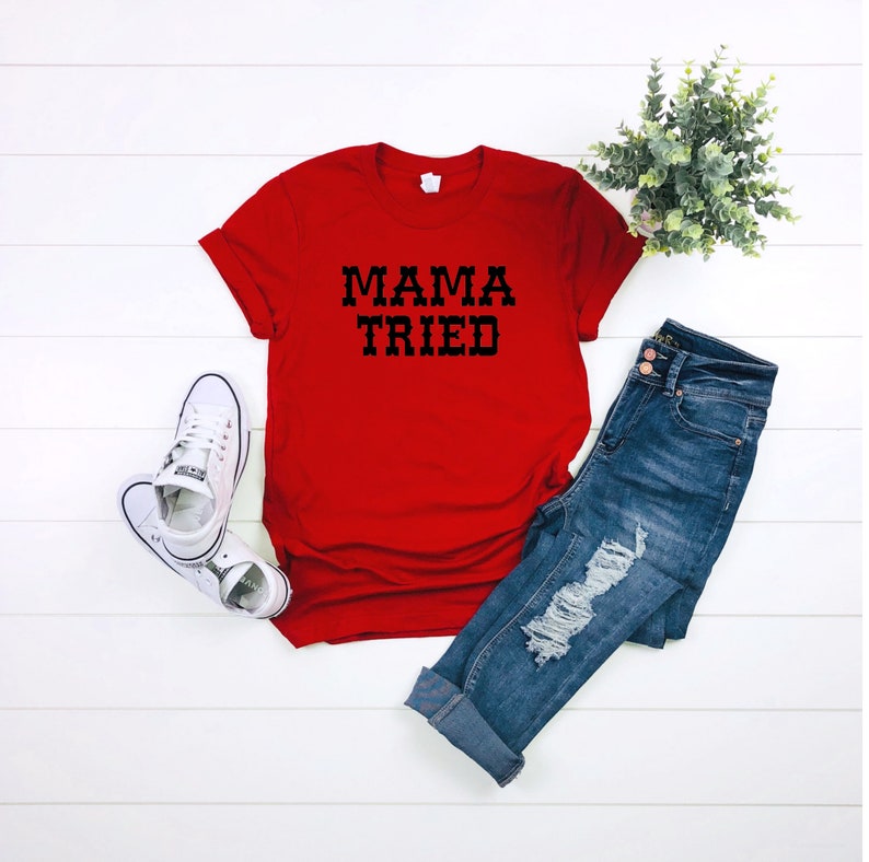 K&ouml;nnte beinhalten: Rotes T-Shirt mit schwarzem Text, der "MAMA TRIED" lautet. Das Shirt liegt auf einer wei&szlig;en Holzoberfl&auml;che mit einem Paar wei&szlig;en Turnschuhen und einer blauen Jeans.