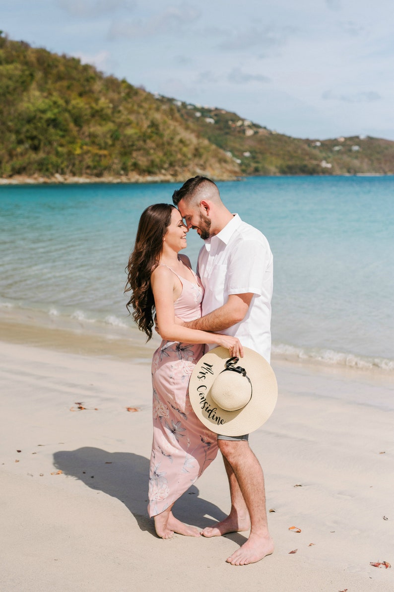 May include: A couple on a beach, the woman is holding a straw hat with the text "Mrs. Considine" written on it. They are looking at each other and smiling.