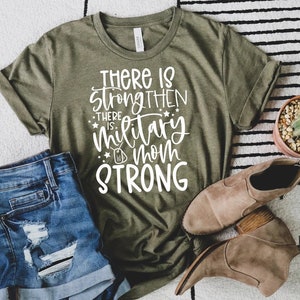 May include: A green t-shirt with white text that reads "There is strength then there is military mom strong". The shirt is laying on a white and black patterned rug with a pair of brown ankle boots and a potted cactus.