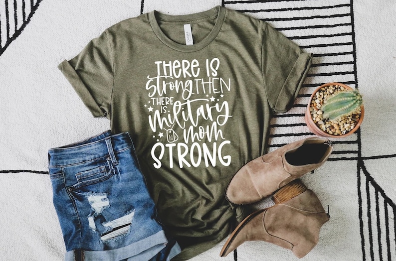 May include: A green t-shirt with white text that reads "There is strength then there is military mom strong". The shirt is laying on a white and black patterned rug with a pair of brown ankle boots and a potted cactus.
