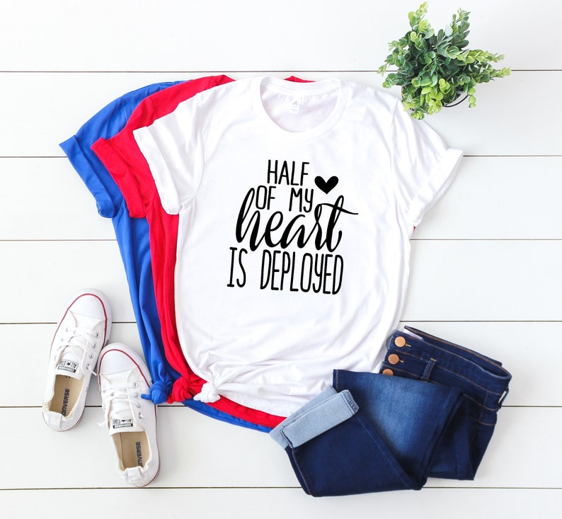 K&ouml;nnte beinhalten: Ein wei&szlig;es T-Shirt mit dem Text "HALF OF MY HEART IS DEPLOYED" in schwarzer Schrift, einem schwarzen Herz und einem blau, rot und wei&szlig;en Farbschema. Das Shirt wird mit blauen Jeans und wei&szlig;en Turnschuhen kombiniert. Eine kleine gr&uuml;ne Pflanze ist im Hintergrund.