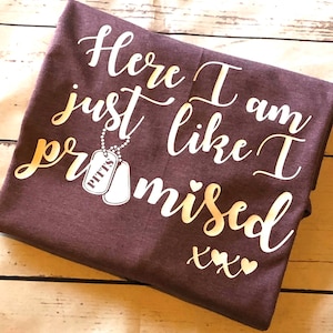 May include: A brown t-shirt with the text "Here I am just like I promised xxx" in gold lettering. A small gold dog tag with the word "Pitts" is attached to a chain above the word "promised".