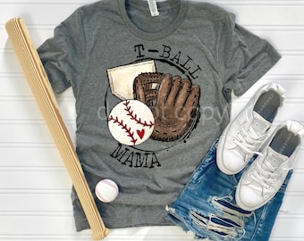 May include: A gray t-shirt with a graphic of a baseball, glove, and bat with the text "T-Ball Mama". The shirt is laid out on a white surface with a wooden baseball bat, a white baseball, and a pair of white sneakers.