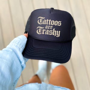 May include: Navy blue trucker hat with the text "Tattoos are Trashy" in a vintage font. The hat has a mesh back and a curved brim. A stylish accessory for anyone who loves tattoos.