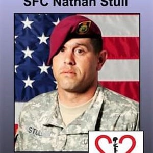 May include: A black and white photo of a soldier in uniform with a red beret. The soldier is standing in front of an American flag. The text "In Memory of SFC Nathan Stull" is at the top of the image. The text "Our Heroes 22 Too Many" is at the bottom of the image. There is a red logo with the number 22 and the text "Too Many" below it.