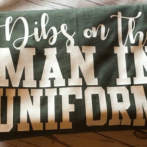 May include: A green t-shirt with white text that reads "Dibs on the Man in Uniform".