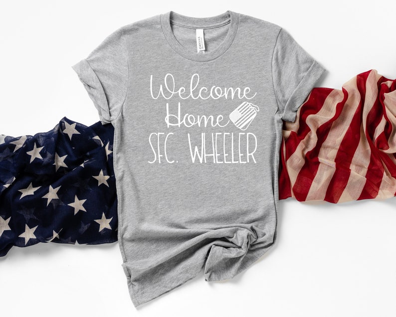 May include: A gray t-shirt with the text "Welcome home SFC. WHEELER" in white lettering. A small white dog tag with an American flag design is next to the text. The t-shirt is displayed on a white background with an American flag scarf on the right side of the image.