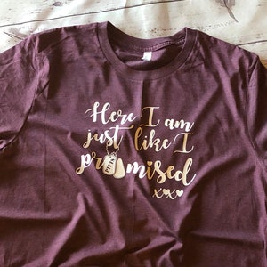 May include: Burgundy t-shirt with the text "Here I am just like I promised xxx" in gold lettering. Two gold dog tags with the word "HITT" on them are hanging from a chain above the text.