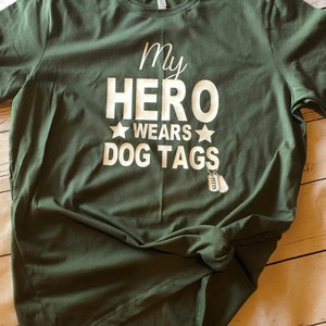 May include: Green t-shirt with white text that reads "My Hero Wears Dog Tags" with a small silver dog tag charm.