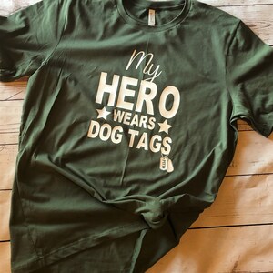 May include: A green t-shirt with the text "My Hero Wears Dog Tags" in white lettering. Two silver dog tags are hanging from the bottom of the text.