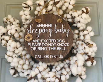 May include: A brown wooden sign with white text that reads "Shhh! Sleeping baby and excited dog please do not knock or ring the bell. Call or text us." The sign is decorated with a cotton wreath.