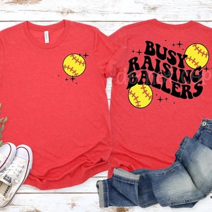 May include: Two red t-shirts with a softball design. One shirt says "Busy Raising Ballers" with a softball graphic. The other shirt has a single softball graphic.