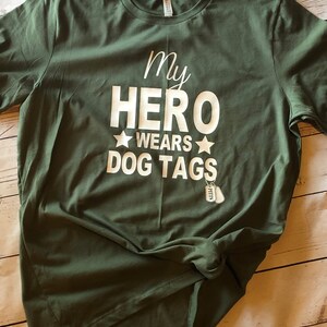May include: A green t-shirt with the text "My Hero Wears Dog Tags" in white lettering. Two small silver dog tags are hanging from the bottom of the text.