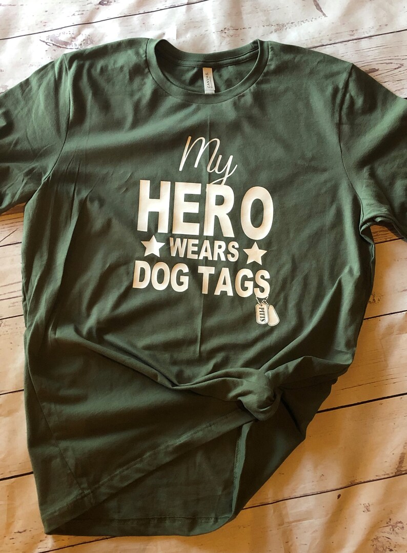 May include: A green t-shirt with the text "My Hero Wears Dog Tags" in white lettering. Two small silver dog tags are hanging from the bottom of the text.