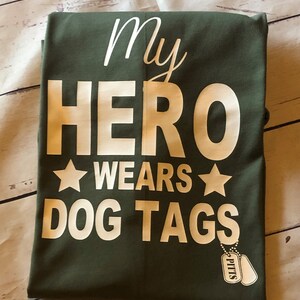 May include: A green t-shirt with white text that reads "My Hero Wears Dog Tags" and a white star on either side of the word "Wears". There are two silver dog tags hanging from the bottom of the shirt that read "Pitts".