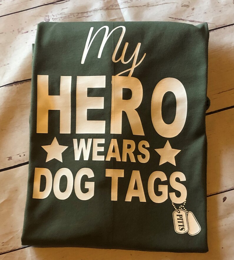 May include: A green t-shirt with white text that reads "My Hero Wears Dog Tags" and a white star on either side of the word "Wears". There are two silver dog tags hanging from the bottom of the shirt that read "Pitts".
