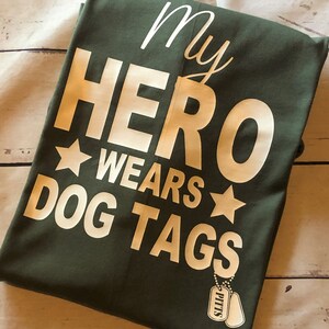 May include: Green t-shirt with white text that reads "My Hero Wears Dog Tags" and a white star on either side of the word "Wears". A small white dog tag with the word "PITTS" is visible in the bottom right corner.