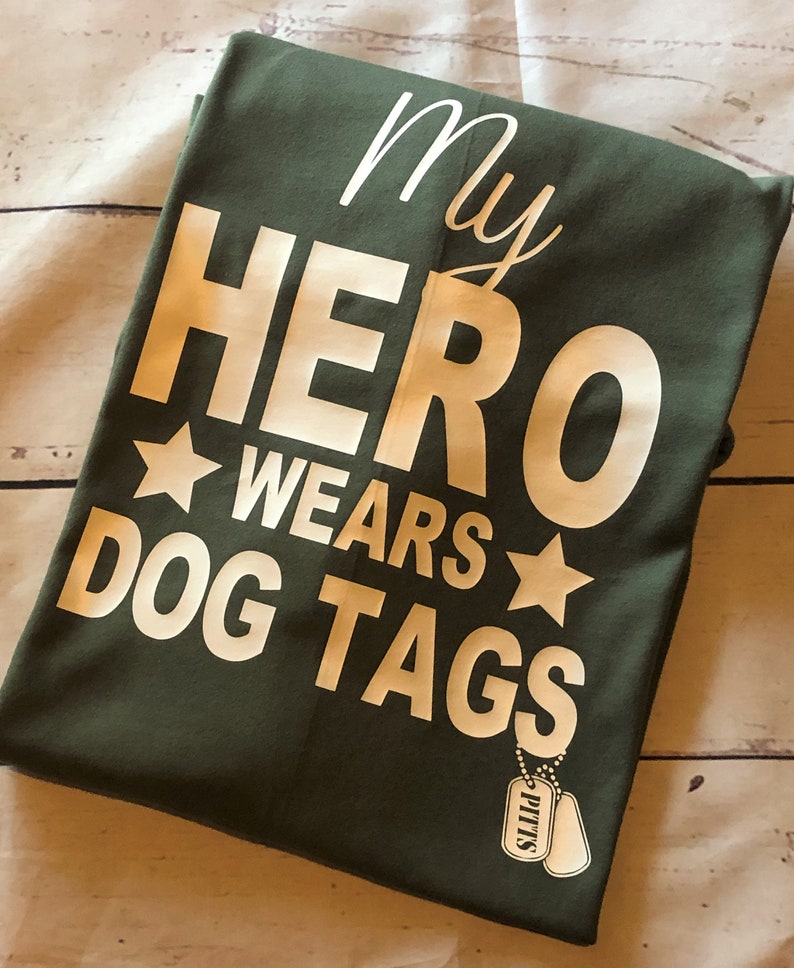 May include: Green t-shirt with white text that reads "My Hero Wears Dog Tags" and a white star on either side of the word "Wears". A small white dog tag with the word "PITTS" is visible in the bottom right corner.