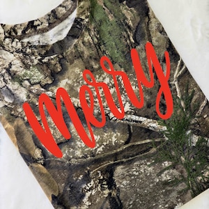 May include: Camouflage-patterned t-shirt with the word "merry" in bright red script across the front. The shirt features a crew neck and a blend of brown, green, and tan colors, creating a woodland camouflage design.