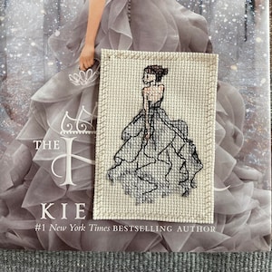 May include: A cross-stitch bookmark depicting a woman in a white gown with a crown on her head. The bookmark is placed on a book with the text "The #1 New York Times Bestselling Author" on the cover.