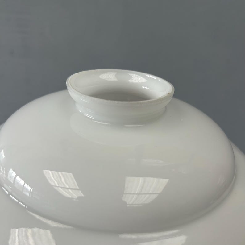 May include: A smooth, white, opaque glass lamp shade with a rounded, dome-like shape. The top features a circular opening with a raised, flared rim designed for mounting. The surface has a glossy finish that reflects light, creating bright highlights.