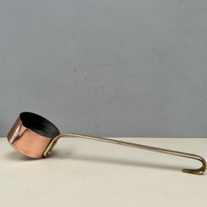 Puede incluir: Un cucharón de cobre con un mango largo de latón. El cuenco del cucharón es de color cobre cálido, y el mango se curva en forma de gancho en el extremo. El cucharón está diseñado para uso en la cocina.