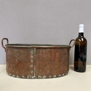 Op de afbeelding: Een grote, antieke koperen kuip met geklonken constructie en twee gebogen handvatten. Het verouderde metaal heeft een rijke, roodbruine patina met groene oxidatie. Een bruine wijnfles met witte dop staat ernaast.