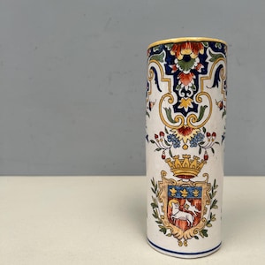 Puede incluir: Un jarrón cilíndrico de cerámica con base blanca, adornado con un diseño floral y heráldico colorido. El jarrón presenta un borde amarillo y un escudo detallado con una corona y un blasón, mostrando una mezcla de arte y motivos históricos.