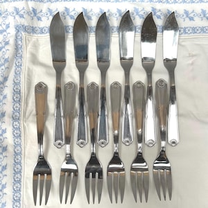 May include: A collection of vintage cutlery, comprising butter knives and forks, displayed on a white cloth with a blue floral pattern. The silverware has a polished, silver appearance.