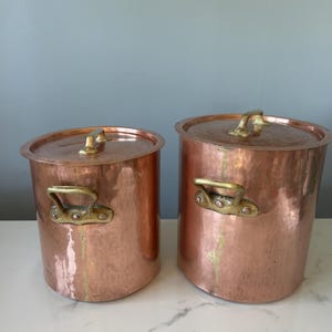 May include: Two cylindrical copper pots with hammered textures, each featuring a flat lid with a small brass handle and two side-mounted brass handles secured by rivets. The pots have a warm metallic finish and show signs of natural patina.