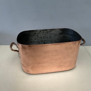 May include: A large, oval-shaped copper pot with two handles. The pot has a dark interior and a shiny, slightly tarnished exterior.